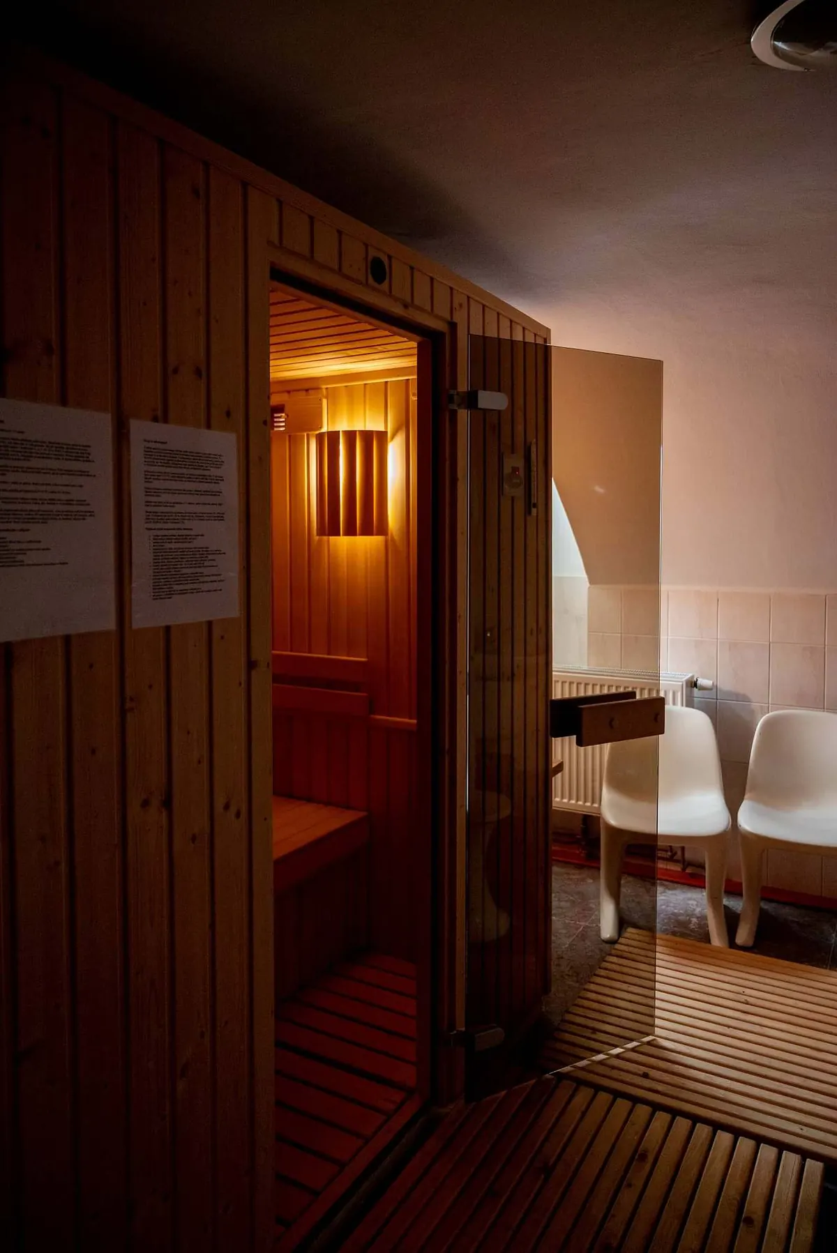 Infrasauna Peklíčko v šeru: otevřené skleněné dveře, uvnitř teplé světlo nad lavicí z dřeva, vpravo bílá křesla odpočívárny