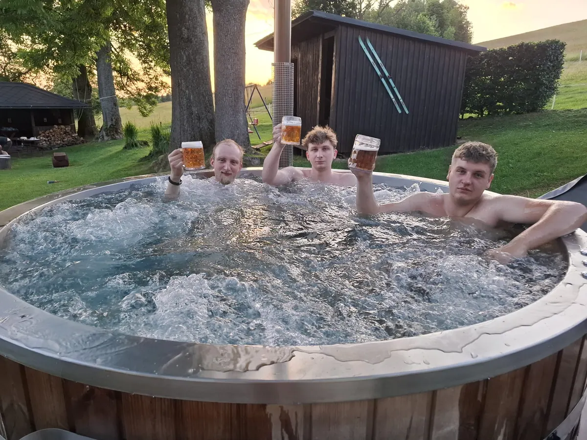 Tři hosté v bublajícím nerezovém sudu s půllitry piva, za nimi zahrada a podvečerní nebe
