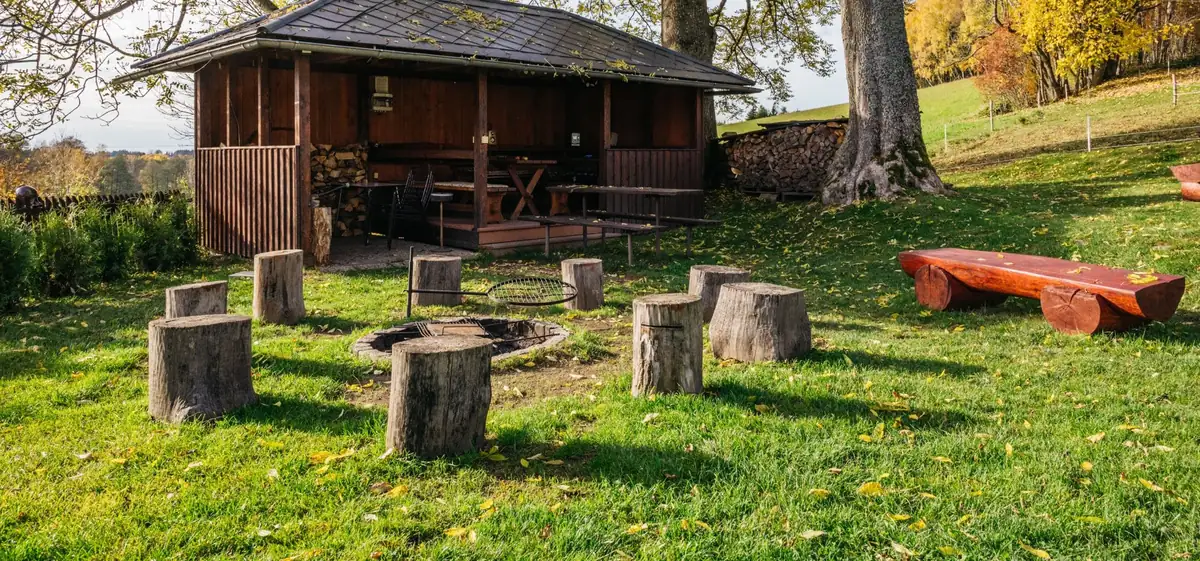 Ohniště s roštem uprostřed pařezů, vpravo lavice z kulatiny, za ohništěm krytý altán s posezením