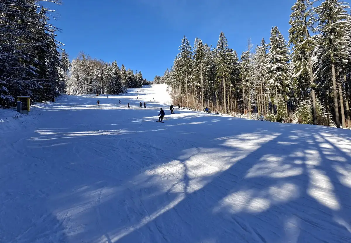 Široká sjezdovka mezi zasněženými smrky za slunečného dne, na svahu lyžaři a snowboardista