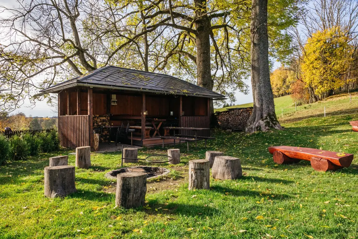 Krytý altán s posezením, ohniště s pařezy a lavice z kulatiny pod vzrostlými stromy