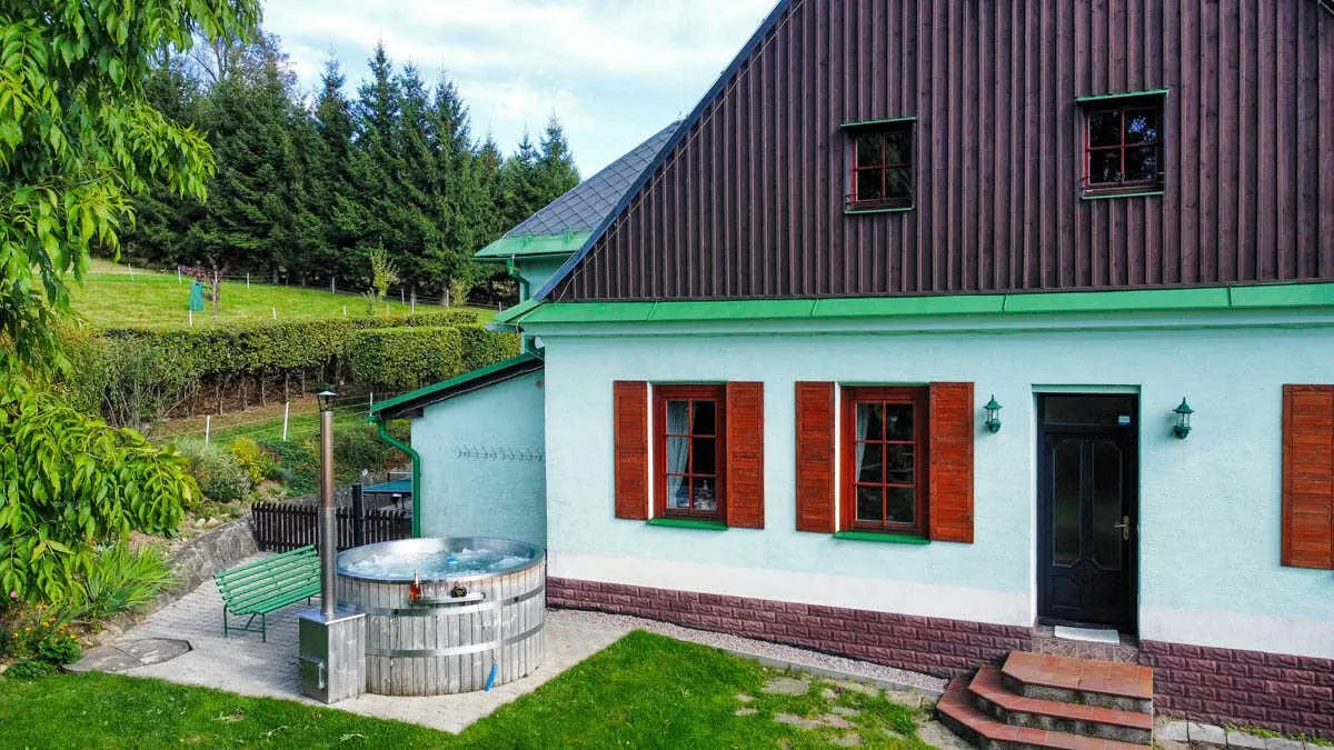 Zahradní strana Severky v létě: mátová fasáda s rezavými okenicemi, vstupní dveře se schody a nerezový koupací sud s kamny na dřevo u zelené lavičky