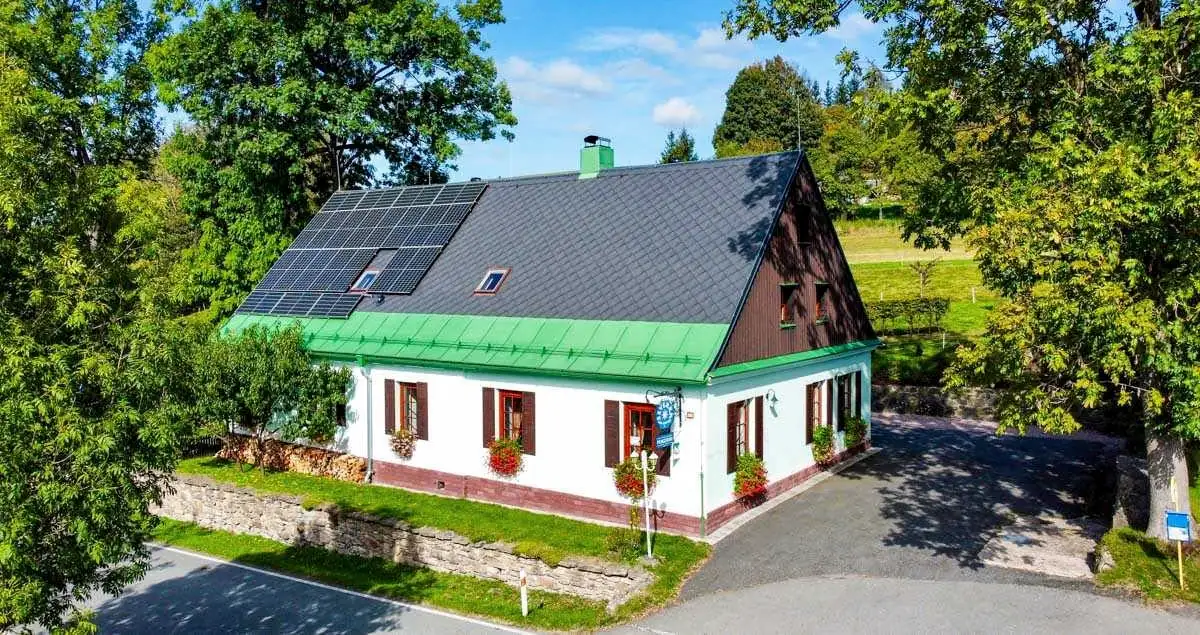 Chata Severka v létě shora od silnice: zelená střecha se solárními panely, mátové přízemí s truhlíky muškátů a kamenná zídka u cesty
